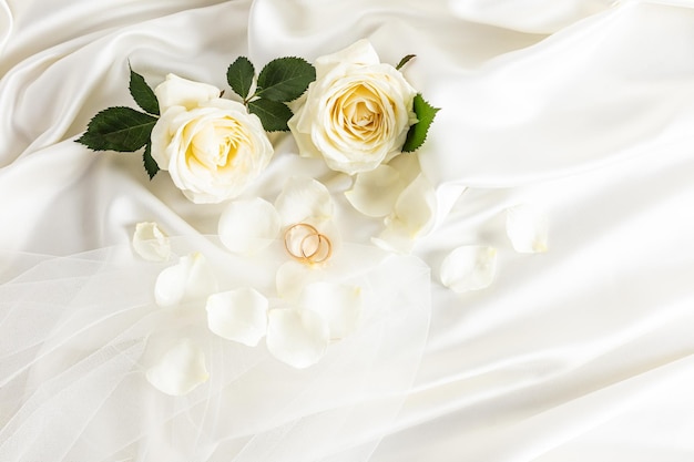 Dos anillos de bodas de oro en un fondo de satén beige con rosas de té vivas y parte del velo de la novia Diseño elegante para el fondo de la boda