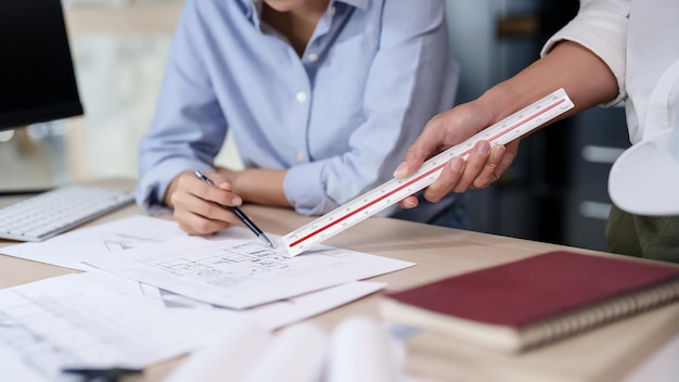 Foto dos arquitectos e ingenieros asiáticos discuten sobre dibujar un proyecto de plan de arquitectura en un plano mientras trabajan en el diseño de un nuevo proyecto sobre el modelo de edificio y el plan arquitectónico de construcción en la oficina