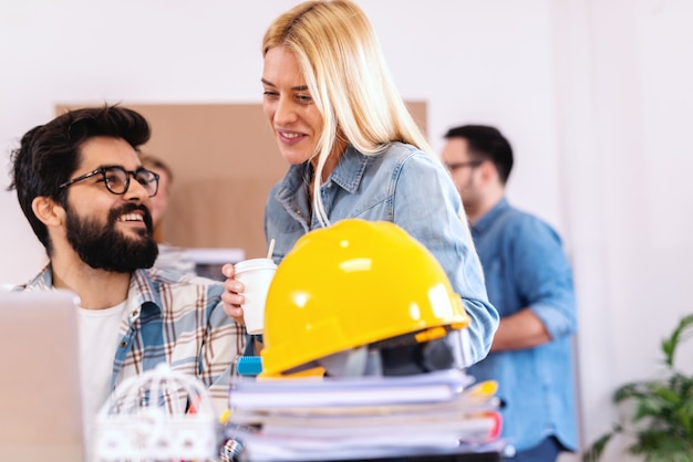 Dos arquitectos mirando portátil y objeto en desarrollo. Anuncio sonriente de la mujer rubia con el café para llevar mientras el hombre barbudo está sentado y mirándola. Poner en marcha el concepto de negocio.