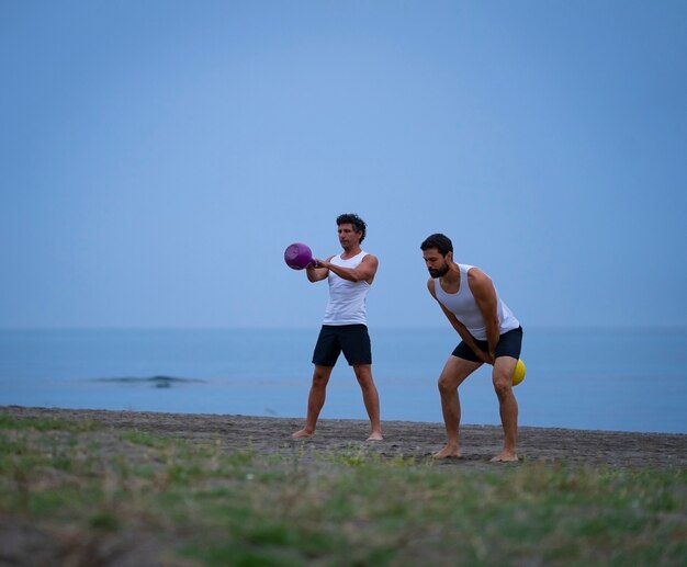 Dos atletas masculinos haciendo ejercicio en la playa con kettle bells