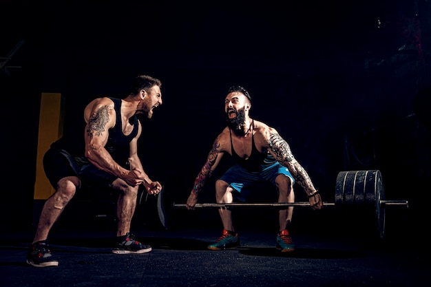 Dos atletas tatuados barbudos musculosos entrenando, uno levanta la barra de peso pesado cuando el otro es motivador