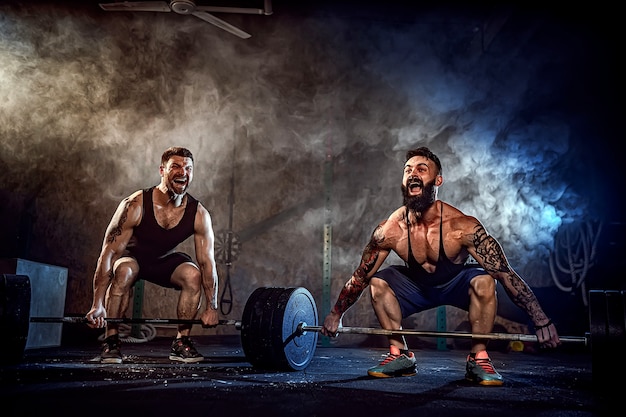 Dos atletas tatuados barbudos musculosos entrenando, levantando la barra de peso pesado en humo en el gimnasio