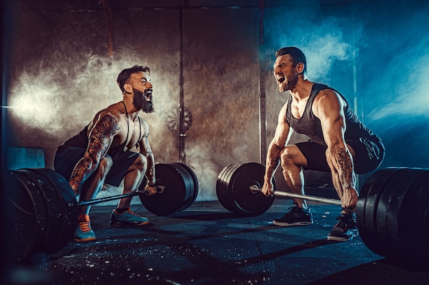 Dos atletas tatuados barbudos musculosos entrenando, levantando la barra de peso pesado en humo en el gimnasio