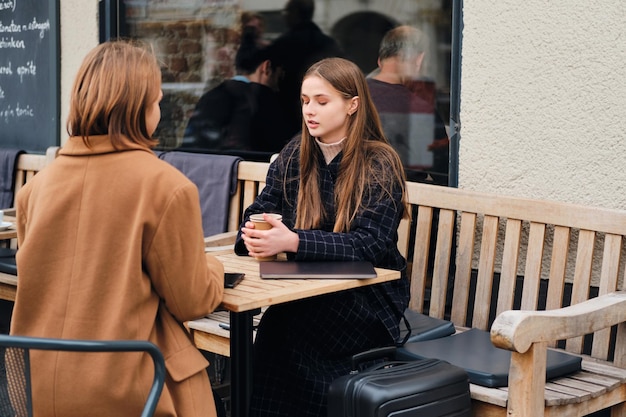 Dos atractivas chicas casuales tomando café juntas mientras están sentadas en un café al aire libre con una maleta