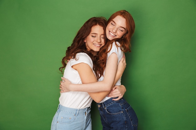 Dos atractivas chicas de jengibre de 20 años en ropa casual sonriendo y abrazándose con los ojos cerrados, aislados sobre fondo verde