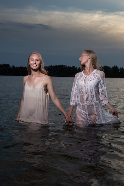 Dos atractivas hermanas gemelas jóvenes con cabello largo y rubio posando en vestidos ligeros en el agua del lago en la noche de verano.