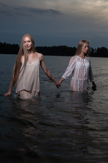 Dos atractivas hermanas gemelas jóvenes con largo cabello rubio posando en vestidos ligeros en el agua del lago por la noche