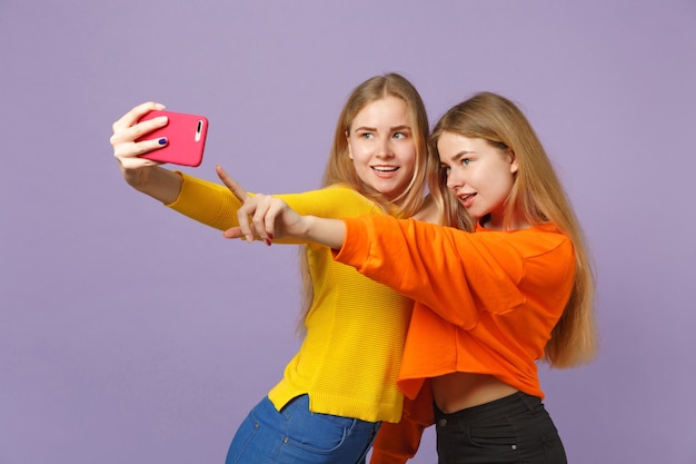 Dos atractivas jóvenes rubias gemelas hermanas niñas en ropa vívida haciendo selfie en teléfono móvil aislado en la pared azul violeta pastel. Concepto de estilo de vida familiar de personas.