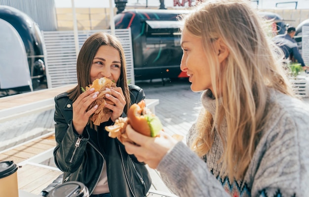 Foto dos atractivas mujeres europeas comen comida rápida en la calle mientras están sentadas en una mesa afuera