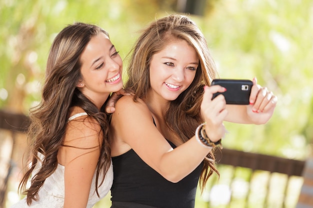 Dos atractivas novias de raza mixta usando sus teléfonos móviles inteligentes para hacer selfies al aire libre