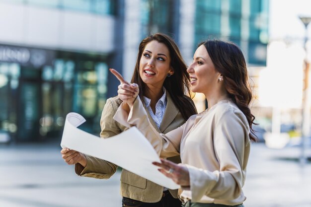 Dos atractivos ingenieros de negocios sonrientes charlando y mirando planes fuera de una oficina.