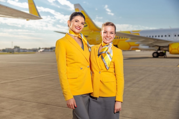 Dos azafatas alegres mirando a la cámara y sonriendo mientras están de pie en el aeródromo con un avión en el fondo