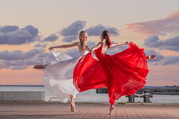 Dos bailarinas con una falda voladora blanca y roja y un leotardo bailando a dúo en el terraplén del océano o el mar contra el telón de fondo del cielo del atardecer