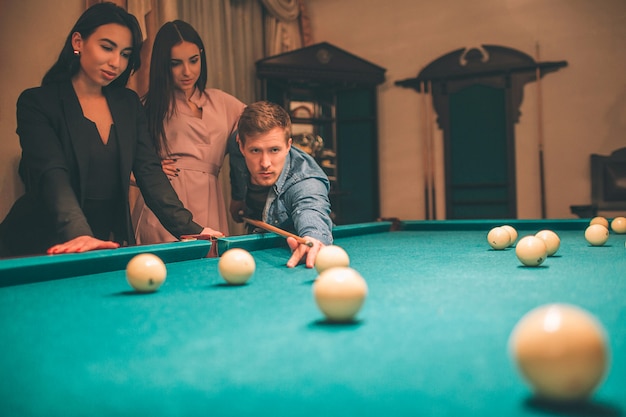 Dos bellas modelos observan cómo el hombre pelirrojo practica billar. Se puso de pie en ángulo con taco de billar y con el objetivo de romper la pelota. Estan en la habitacion.