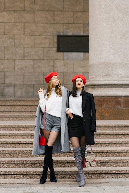 Dos bellas mujeres francesas con boinas, mirando a la cámara y sonriendo mientras caminan juntas al aire libre cerca de los escalones de un edificio clásico.