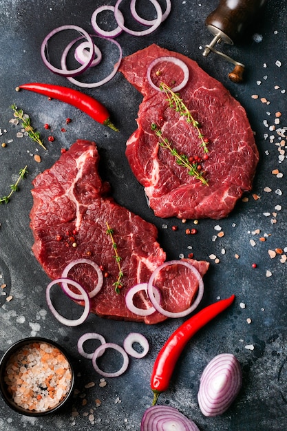 Dos bistec de ternera cruda con especias, cebollas y chile sobre fondo de hormigón o pizarra oscura. Vista superior