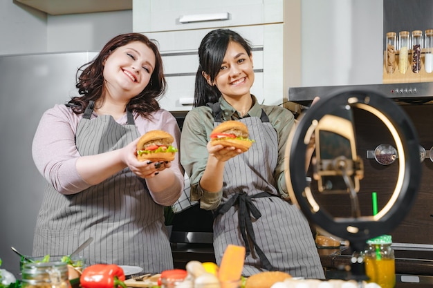 Dos blogueras sonrientes preparando hamburguesas y grabando videos en un teléfono inteligente para redes sociales o un blog en la cocina.