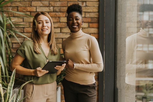 Dos bonitas mujeres de negocios jóvenes con tableta digital de pie junto a la pared de ladrillo en la oficina de estilo industrial