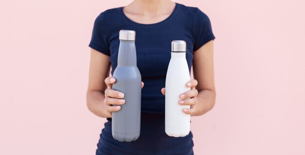 Dos botellas de agua termo de acero reutilizables en manos femeninas en el fondo de color rosa pastel.