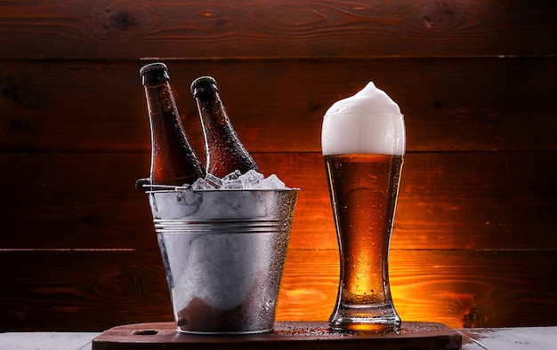 dos botellas de cerveza en un cubo con hielo y un vaso de cerveza con espuma exuberante