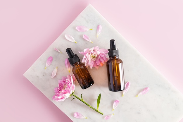 Dos botellas cosméticas de vidrio oscuro con un producto orgánico natural se encuentran sobre baldosas de mármol blanco entre las flores y los pétalos de fondo rosa de primera vista