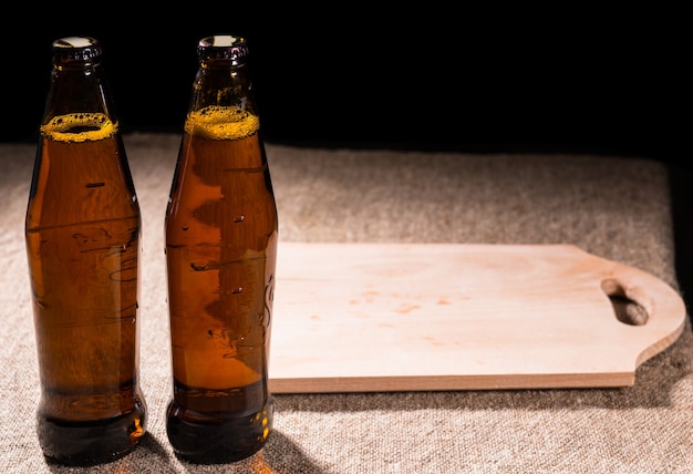 Dos botellas llenas de cerveza en una mesa cubierta de arpillera junto a una tabla de cortar de madera rústica