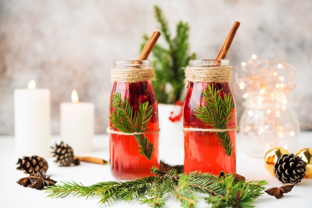 Dos botellas de vino caliente caliente con bayas y especias están en el blanco con luces navideñas y decoraciones navideñas.