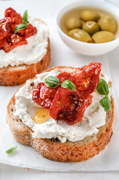 Dos bruschettas con aceitunas de queso ricotta y tomates secos en pan ciabatta sobre fondo blanco.