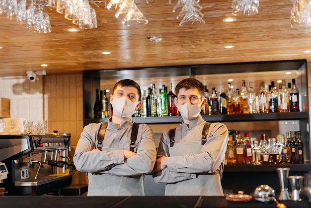 Dos camareros elegantes con máscaras y uniformes durante la pandemia se paran detrás de la barra El trabajo de los restaurantes y cafés durante la pandemia