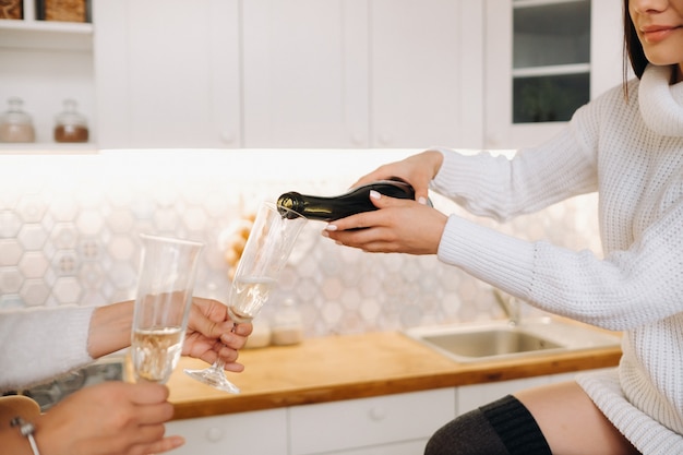 Dos chicas en un acogedor ambiente hogareño en la cocina vierten champán para Navidad. Chicas sonrientes beben champán en una noche festiva