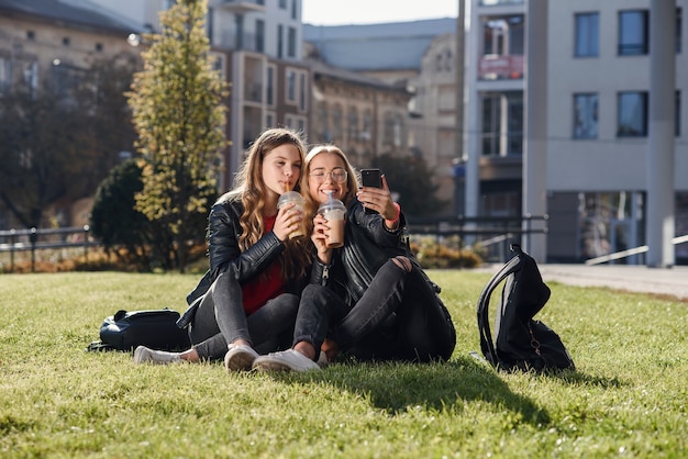 Dos chicas adolescentes atractivas con estilo con batido y smartphone sentados en el césped. Tiempo libre con mejores amigos.