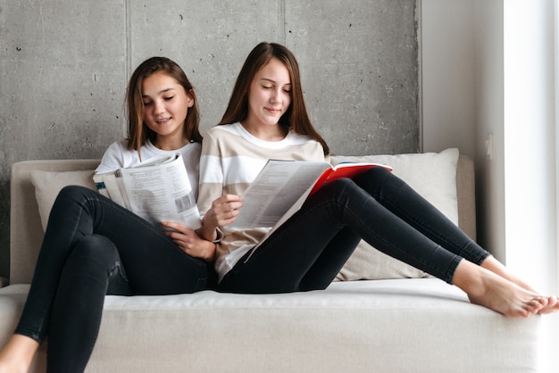 Dos chicas adolescentes sonrientes leyendo revistas mientras están sentados en un sofá en casa