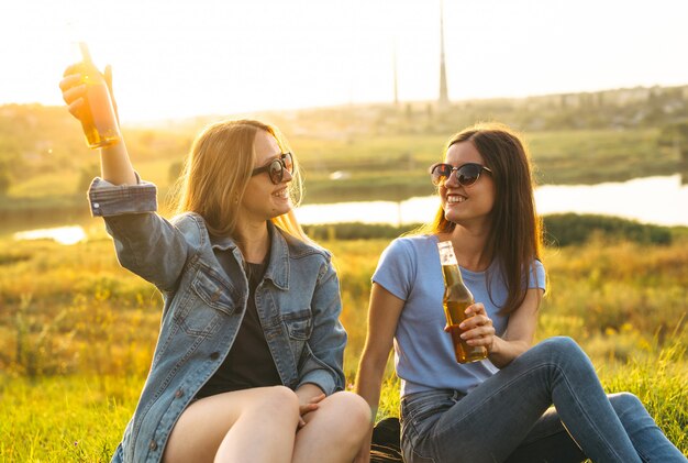 Dos chicas alegres y jóvenes amigos con gafas de sol, bebiendo cerveza y disfrutando del tiempo que pasaron juntos al atardecer.