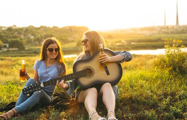 Dos chicas alegres y jóvenes amigos con gafas de sol, bebiendo cerveza y disfrutando del tiempo que pasaron juntos al atardecer.