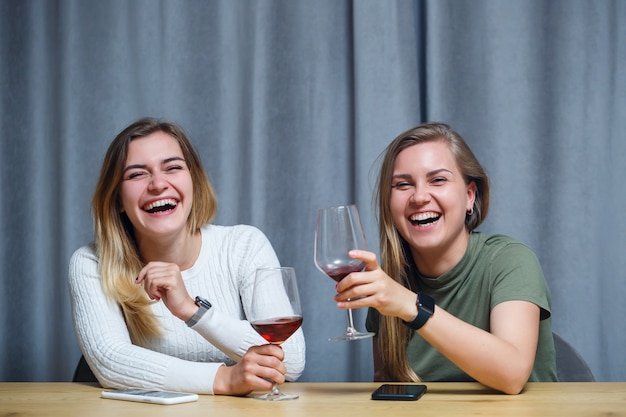 Foto dos chicas de apariencia europea con cabello rubio están sentadas a la mesa, bebiendo vino y riendo, relajándose en casa, alcohol