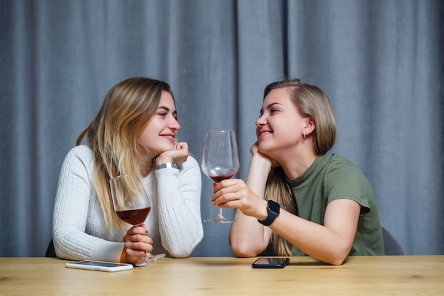 Dos chicas de apariencia europea con cabello rubio están sentadas a la mesa, bebiendo vino y riendo, relajándose en casa, alcohol