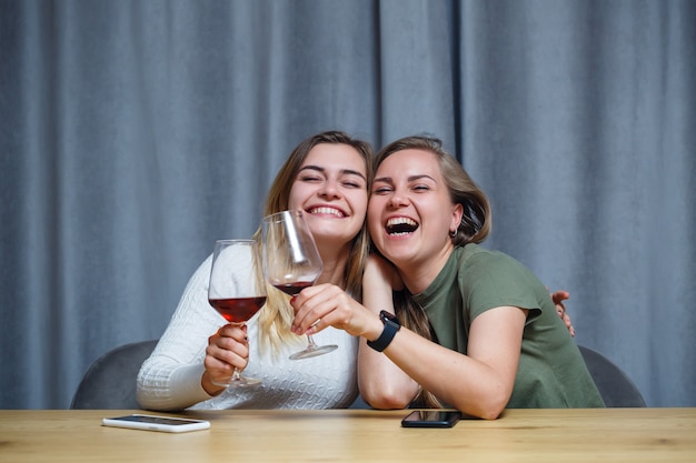 Foto dos chicas de apariencia europea con cabello rubio están sentadas a la mesa, bebiendo vino y riendo, relajándose en casa, alcohol