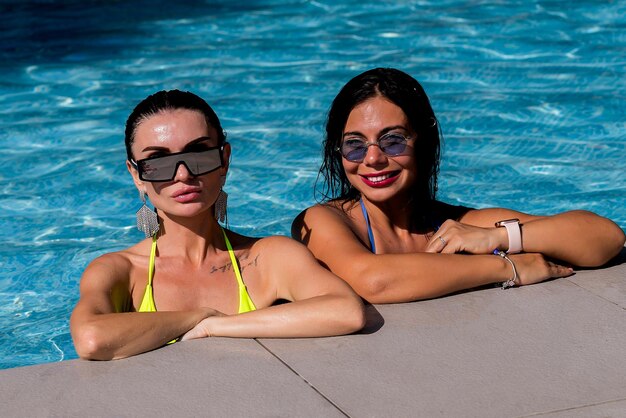 Dos chicas atractivas con cabello largo están posando cerca de la piscina en el sol