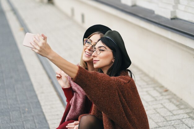 Dos chicas atractivas haciendo selfie sentados juntos, morena y rubia. Traje elegante para la temporada de frío.