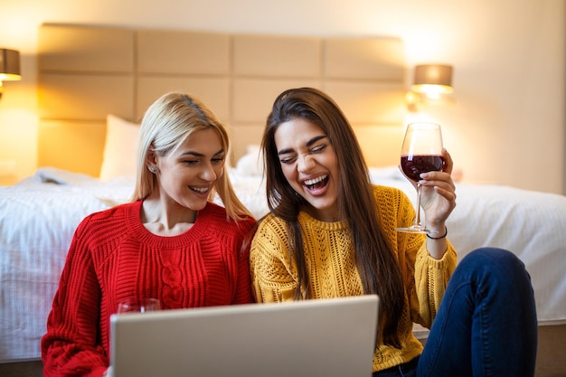 Dos chicas atractivas mejores amigas alegres divirtiéndose viendo películas en la computadora portátil y bebiendo vino rojo en casa dos vasos de vino blanco en las manos