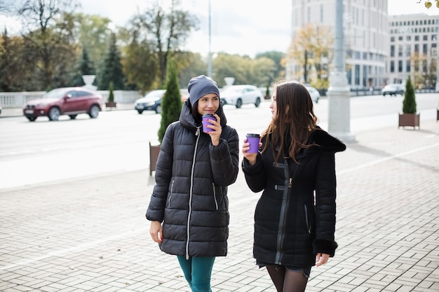 Foto dos chicas caminan por la ciudad hablando, tazas en la mano.