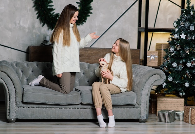 Dos chicas encantadoras posando con expresión de cara de besos con árbol de Navidad. Señora europea de pelo largo de pie junto a árboles y regalos