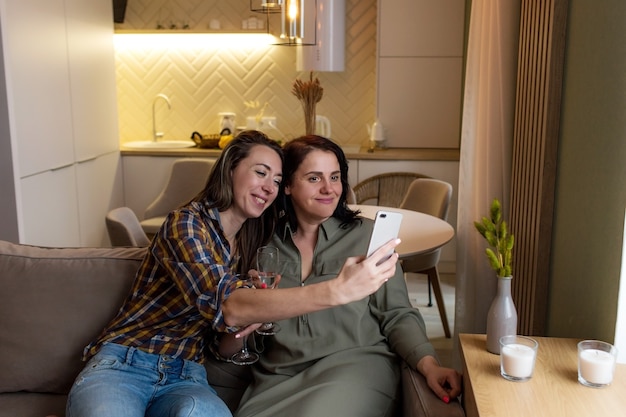 Dos chicas están sentadas en el sofá, levantando sus vasos y tomando selfies en su teléfono.