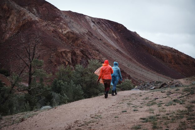 Dos chicas con excelente humor en impermeables viajan. Clima lluvioso en las montañas impide el trekking. Ir por un sendero de montaña.