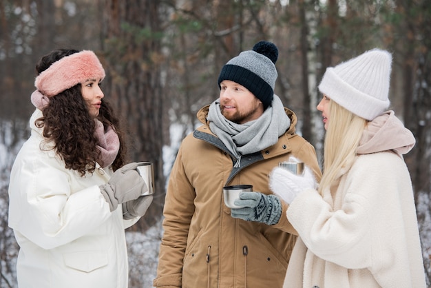 Dos chicas guapas en abrigos blancos de invierno tomando té caliente y hablando con un joven entre árboles en el bosque o parque