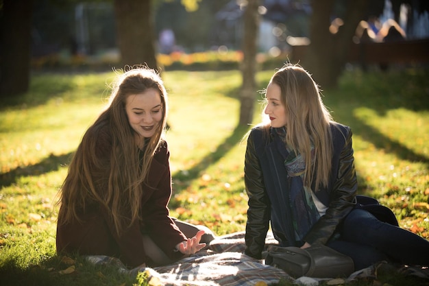 Dos chicas guapas con abrigos hablando sentadas en un plaid en el parque de otoño