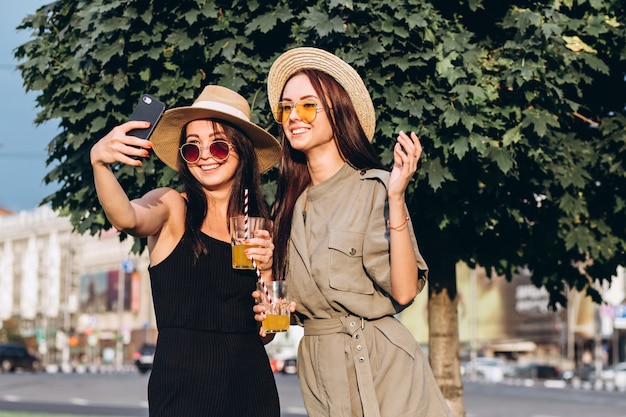 Dos chicas guapas toman selfie en el verano al aire libre al atardecer. Las novias se divierten, ríen, sonríen y toman fotos. Closeup retrato de dos mujeres jóvenes en sombreros de paja.