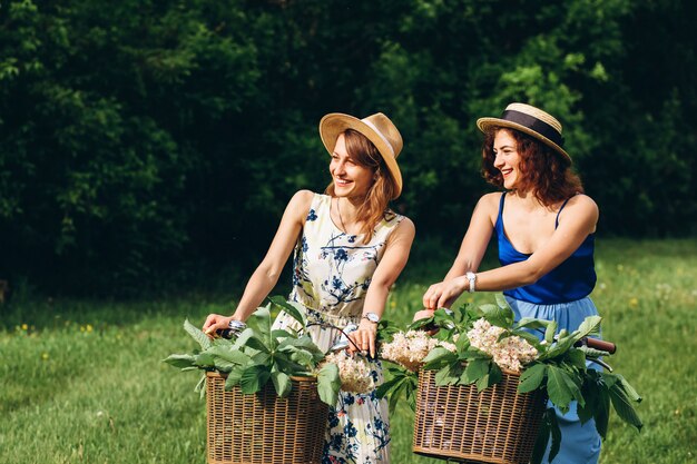 Dos chicas guapas van con bicicletas en un parque de primavera al atardecer. Las novias se divierten, ríen, sonríen y andan en bicicleta. Closeup retrato de dos mujeres jóvenes en vestidos y sombreros de paja. Vacaciones activas de verano