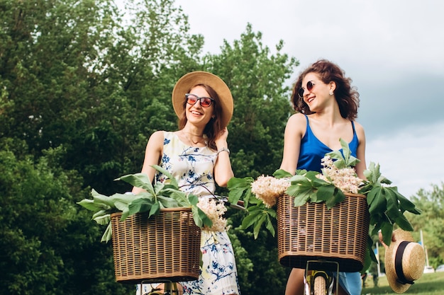 Dos chicas guapas van con bicicletas en un parque de primavera al atardecer. Las novias se divierten, ríen, sonríen y andan en bicicleta. Closeup retrato de dos mujeres jóvenes en vestidos y sombreros de paja. Vacaciones activas de verano