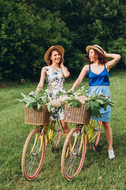 Dos chicas guapas van con bicicletas en el parque de la primavera al atardecer. Las novias se divierten, ríen, sonríen y andan en bicicleta. Retrato de cuerpo entero de dos mujeres jóvenes en vestidos y sombreros de paja. vacaciones activas